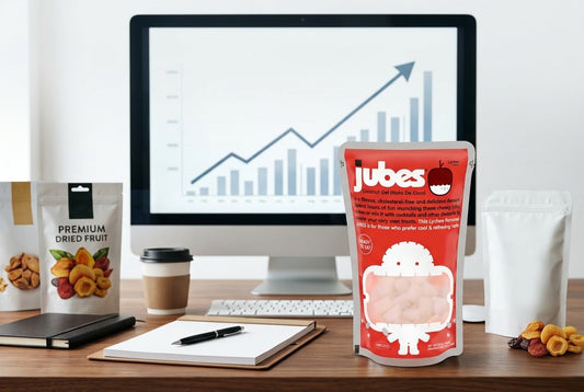 A computer screen showing an upward traffic chart next to a Jubes Lychee pouch, dried fruit bags, a coffee cup, and a notebook on a desk.