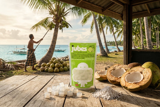 A Jubes Original coconut jelly pouch on a wooden table with halved coconuts, shredded coconut, and a man harvesting coconuts from a palm tree behind it.