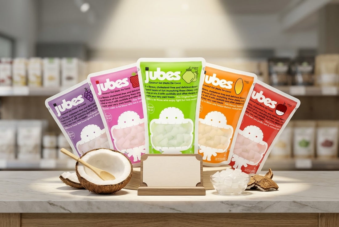 Five Jubes coconut jelly flavor pouches fanned out on a counter with a halved coconut, a bowl of gel cubes, and a blank shelf display sign.