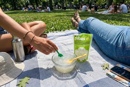 Two people on a picnic blanket share a bowl of Jubes coconut jelly cubes with colorful spoons, a green Jubes pouch and water bottle nearby.