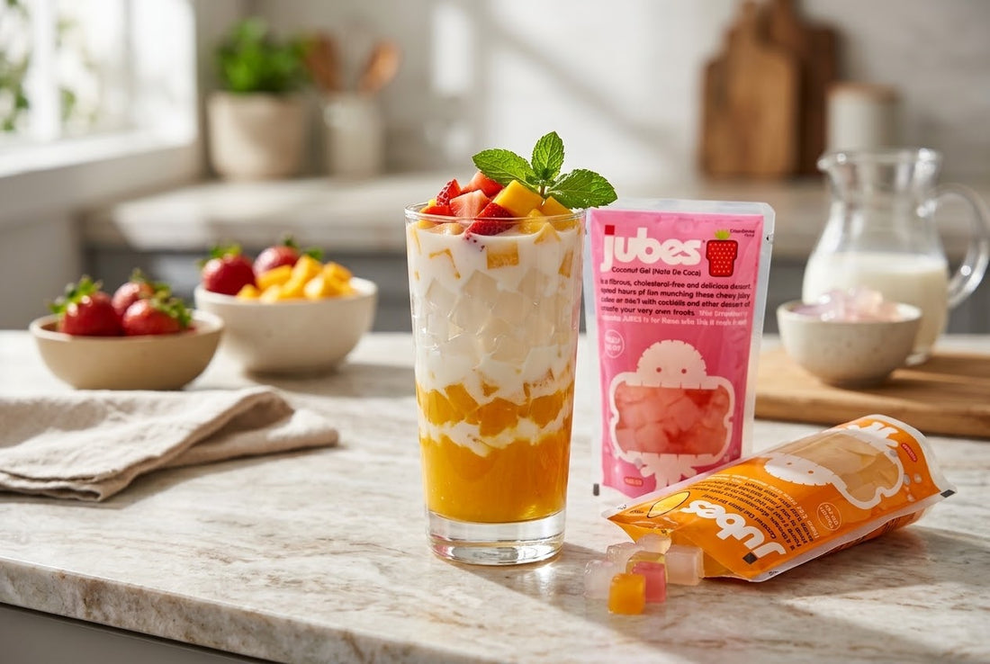 A layered mango and cream drink in a tall glass topped with strawberries and mint, next to Strawberry and Mango Jubes pouches and fresh fruit.