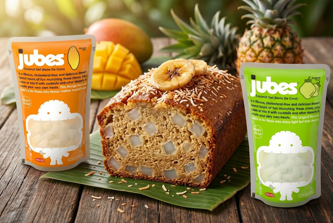 Sliced tropical banana bread with Jubes coconut gel cubes between Jubes Mango and Original pouches, with mango and pineapple behind.