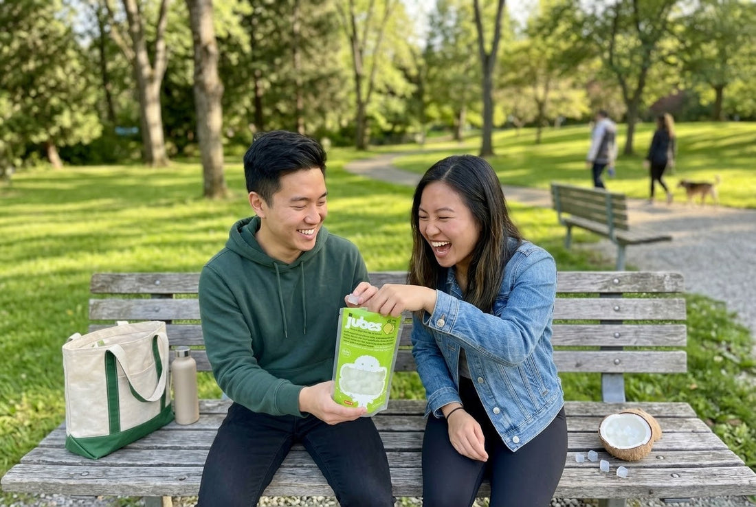 Two friends laughing on a park bench, sharing a green Jubes Nata De Coco pouch, with a coconut half full of jelly cubes beside them.