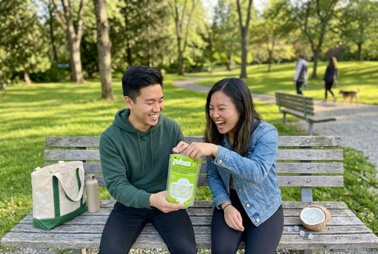 Two friends laughing on a park bench, sharing a green Jubes Nata De Coco pouch, with a coconut half full of jelly cubes beside them.