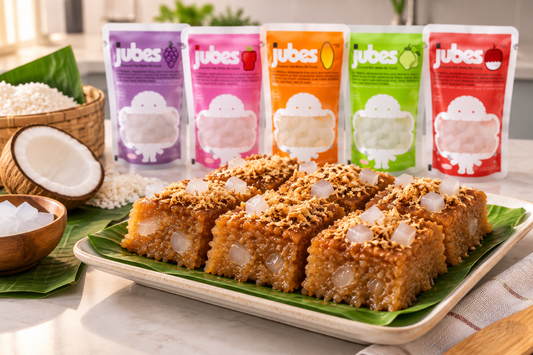 Sliced biko squares topped with latik and jelly cubes on a banana leaf platter, with five Jubes flavor pouches lined up behind, next to a coconut.