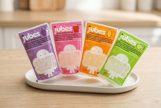 Four Jubes coconut jelly pouches in grape, strawberry, mango, and lychee flavors displayed upright together on a white oval serving tray.
