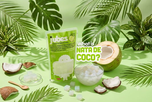 A bag of Jubes Original Nata De Coco beside a bowl of coconut jelly cubes, fresh coconut halves, and green tropical leaves.