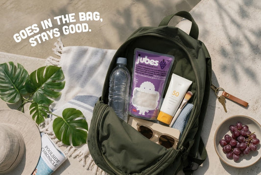 Open backpack packed with Jubes Grape coconut jelly, sunscreen, sunglasses, and water, beside a towel, sunhat, and bowl of grapes.