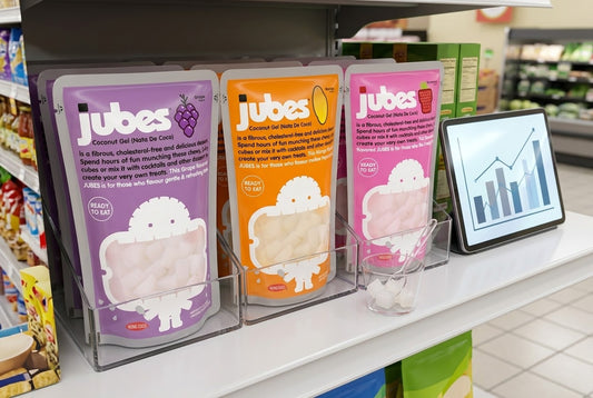 Three Jubes coconut gel pouches, grape, mango, and strawberry, on a grocery shelf display beside a tablet showing an upward performance chart.