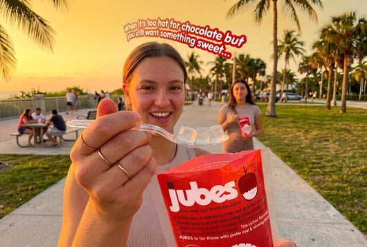 A woman holding a fork of coconut jelly cubes and a red Jubes Lychee pouch at sunset on a palm-lined beach boardwalk.