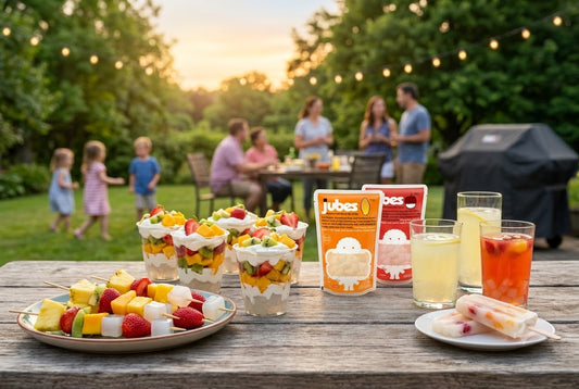 A wooden picnic table filled with fruit parfaits, fruit skewers, popsicles, and refreshing drinks featuring orange and red pouches of Jubes Nata De Coco, while a family gathers in a sunny backyard in the background.