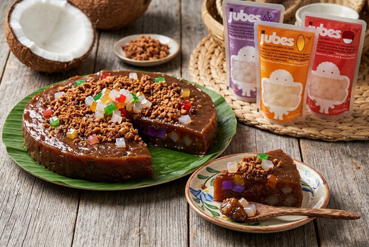 A sliced kalamay coconut rice cake topped with fruit jelly cubes and toasted coconut crumble on a banana leaf, with Jubes pouches behind it.